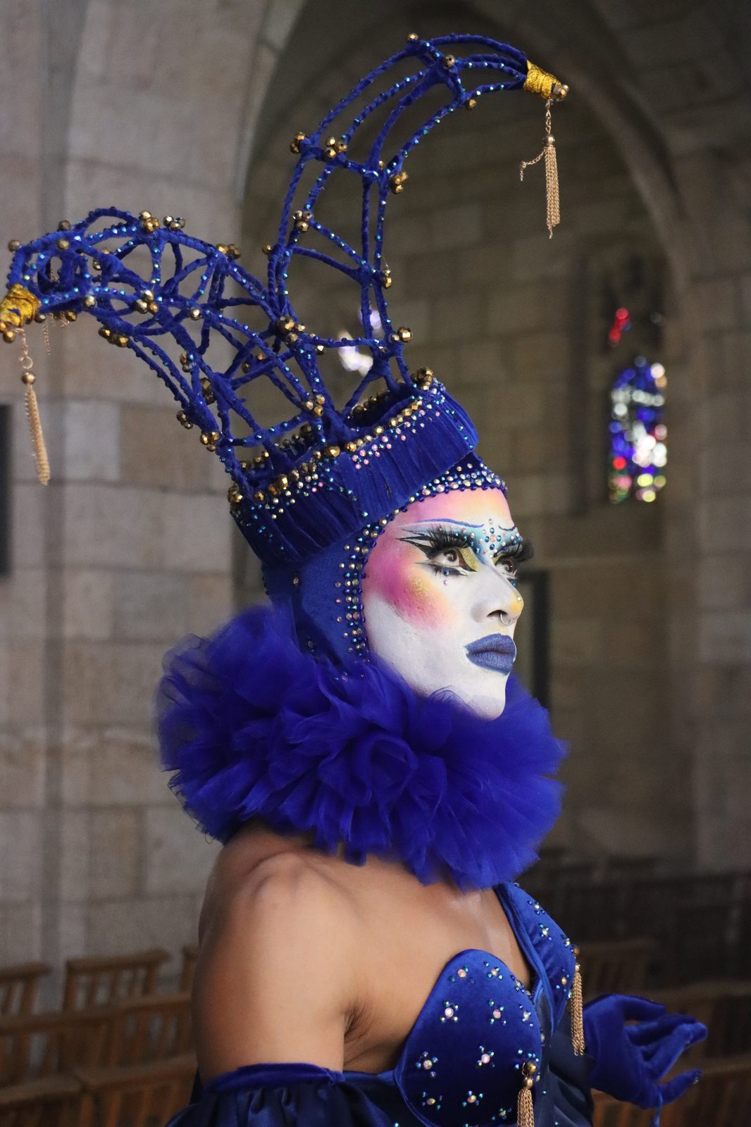 Ina Propriette in an elaborate blue headdress and beaded ruff costume, in profile inside a stone cathedral, from Beaulah: Queens van die Kaap